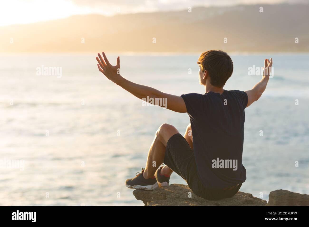 Glücklicher junger Mann, der sich gut fühlt, auf einer Bergklippe mit Blick auf den Ozean zu sitzen und die Arme zum Himmel zu heben. Hoffnung und Dankbarkeit. Stockfoto