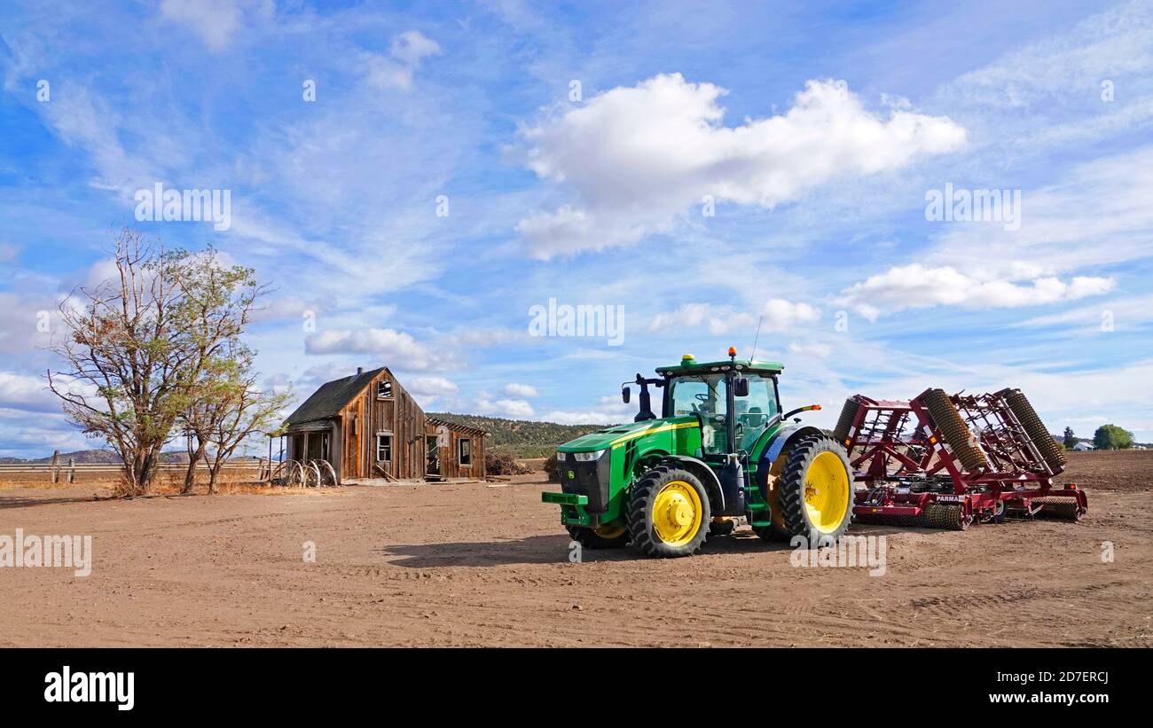 Ein neuer Traktor von John Deere, der einen Scheibenpflug zieht, befindet sich in der Nähe eines verlassenen Bauernhauses aus der Zeit der Depression in der Nähe der kleinen Bauernstadt Culver im Zentrum von Oregon. Stockfoto