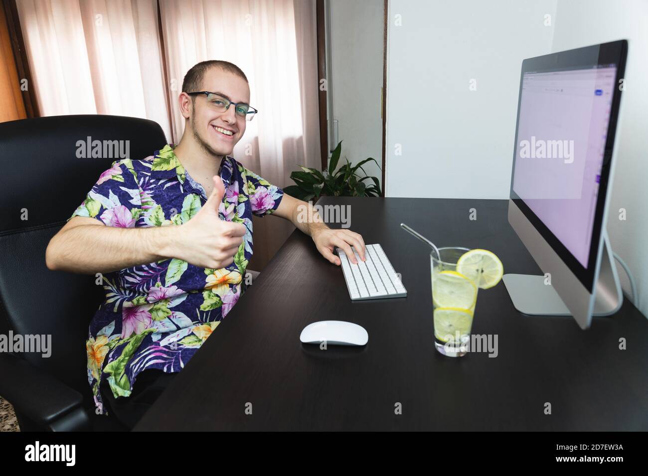 Junger Mann, der am Computer arbeitet, mit hawaiiihemd Fernarbeit, mit einem erfrischenden Getränk. Entspannender Job. Urlaub und Arbeit kombiniert. Lächelnd an Stockfoto