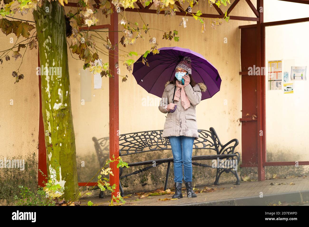 Frau mit Regenschirm an der Bushaltestelle, die auf dem Handy spricht Stockfoto