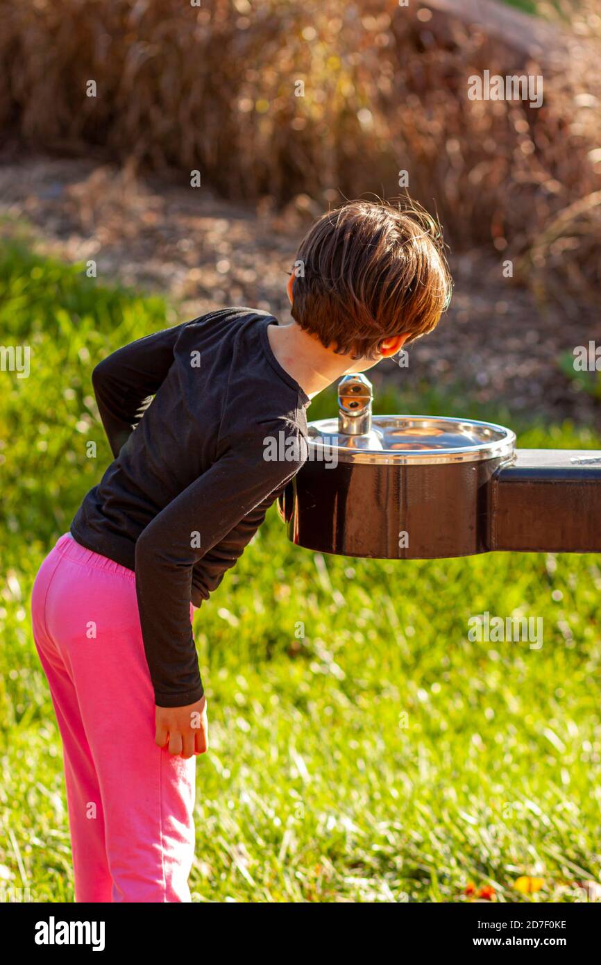 Ein niedliches kleines kaukasisches Mädchen mit kurzen braunen Haaren beugt sich vor, um Wasser aus einem öffentlichen Wasserspender in einem Park zu trinken. Durstige Mädchen drückt die Stockfoto