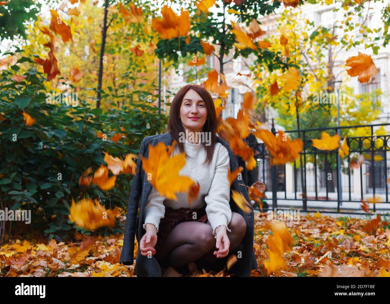Hübsche lächelnde junge Frau wirft gelbe Ahornblätter im öffentlichen Park. Genießen Sie den indischen Sommer. Herbstkonzept. Stockfoto