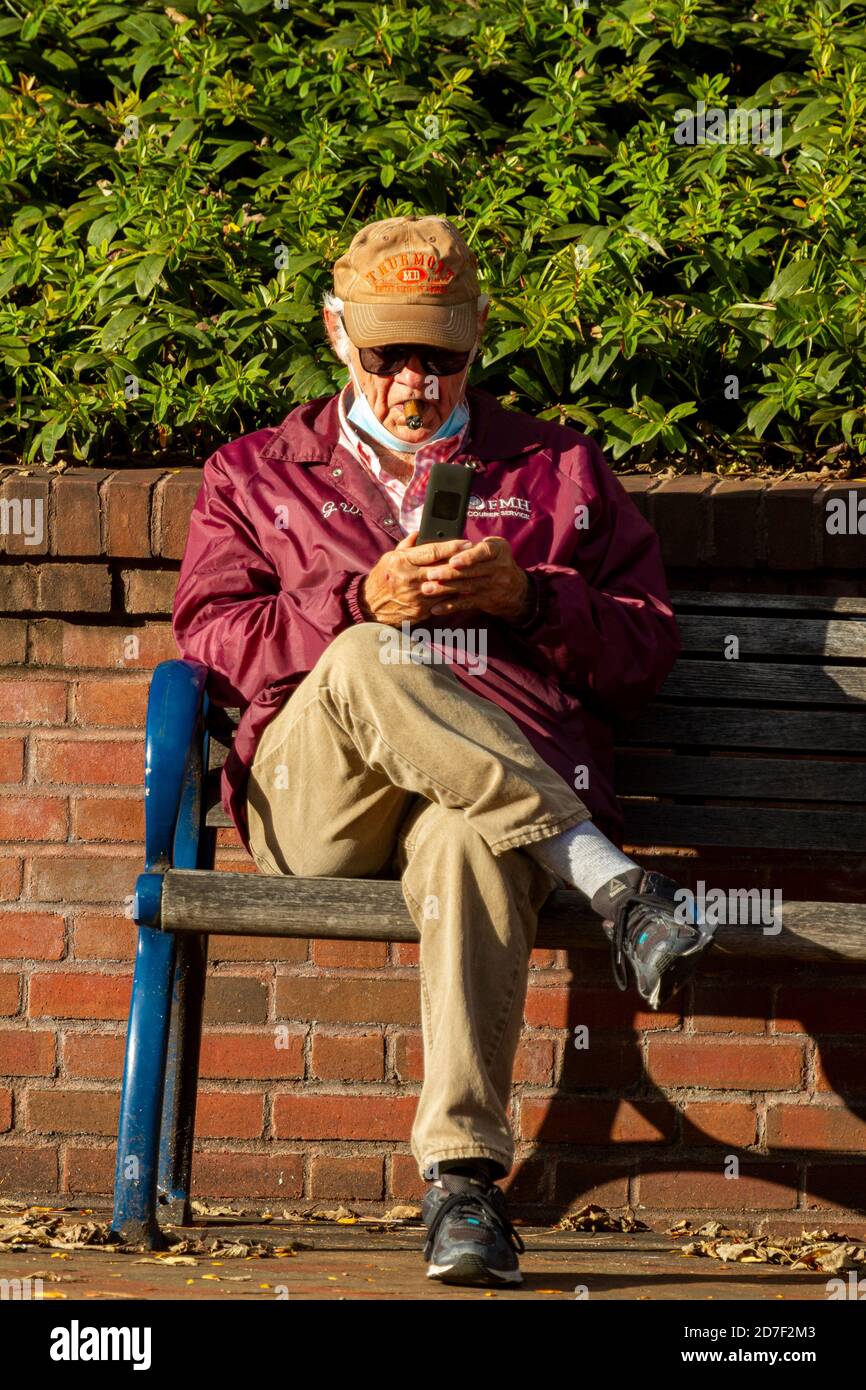 Frederick, MD, USA 10/14/2020: Ein älterer kaukasischer Mann mit Gesichtsmaske sitzt auf einer Parkbank mit einer Zigarre im Mund und versucht es Stockfoto