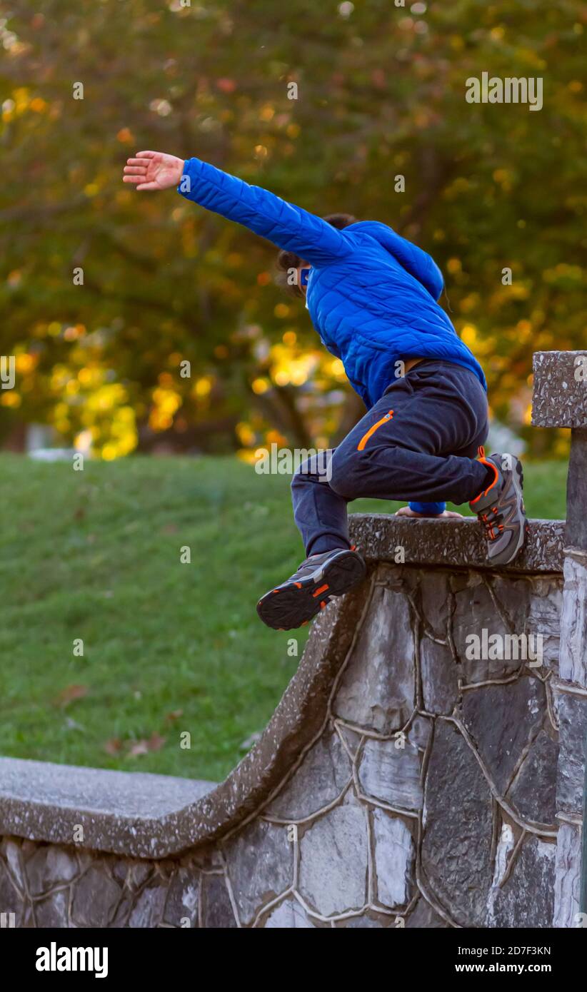 Ein hyperaktives Kind springt von einer hohen Wand, als er wegläuft. Der mutige Junge trägt Track Pants, Sneakers und einen Kapuzenmantel. Er ist voll von en Stockfoto