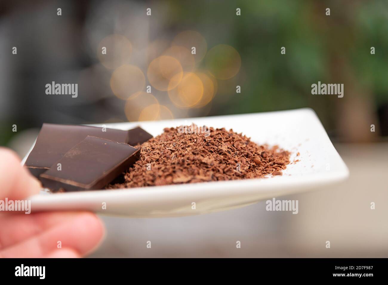 Frauen Hand hält einen weißen Teller. Auf dem Teller ist geriebene Schokolade. Im Hintergrund ist festliches Bokeh. Stockfoto