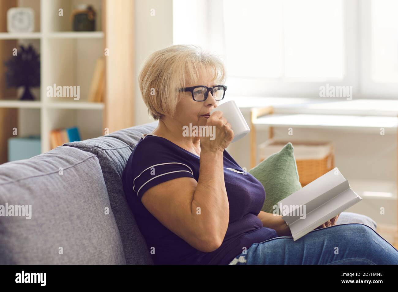 Reife Frau in Gläsern, die sich zu Hause auf der Couch entspannt, Tee trinkt und allein ein Buch liest Stockfoto