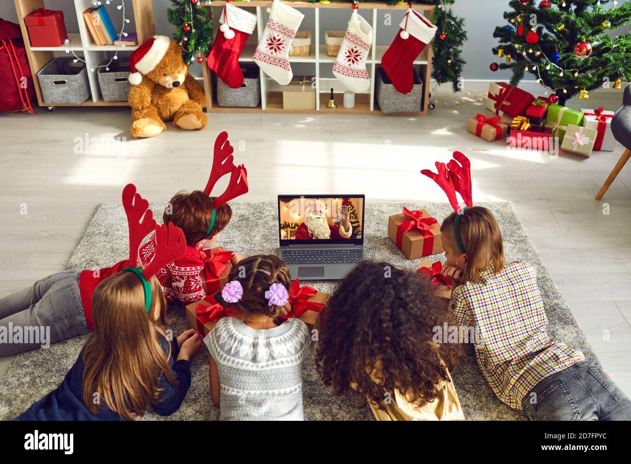 Eine Gruppe von Kindern, die auf dem Boden liegen und einen Video-Chat mit dem Weihnachtsmann mit einem Laptop in einem dekorierten Raum während Weihnachten und Neujahr sehen. Stockfoto