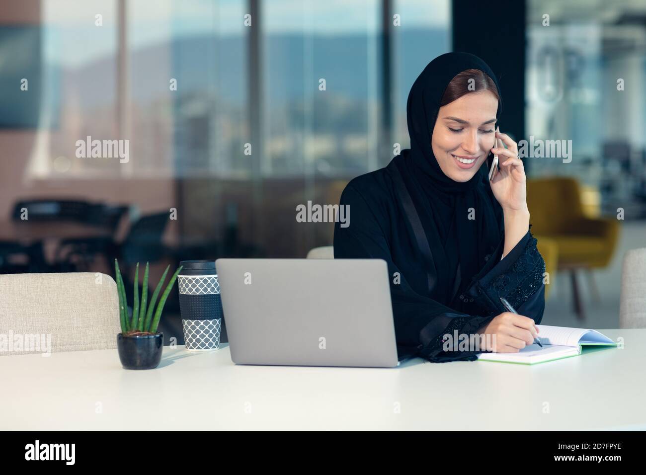 Glückliche muslimische Geschäftsfrau in Hijab am Büroarbeitsplatz. Lächelnde arabische Frau, die am Laptop arbeitet und mit dem Smartphone spricht Stockfoto