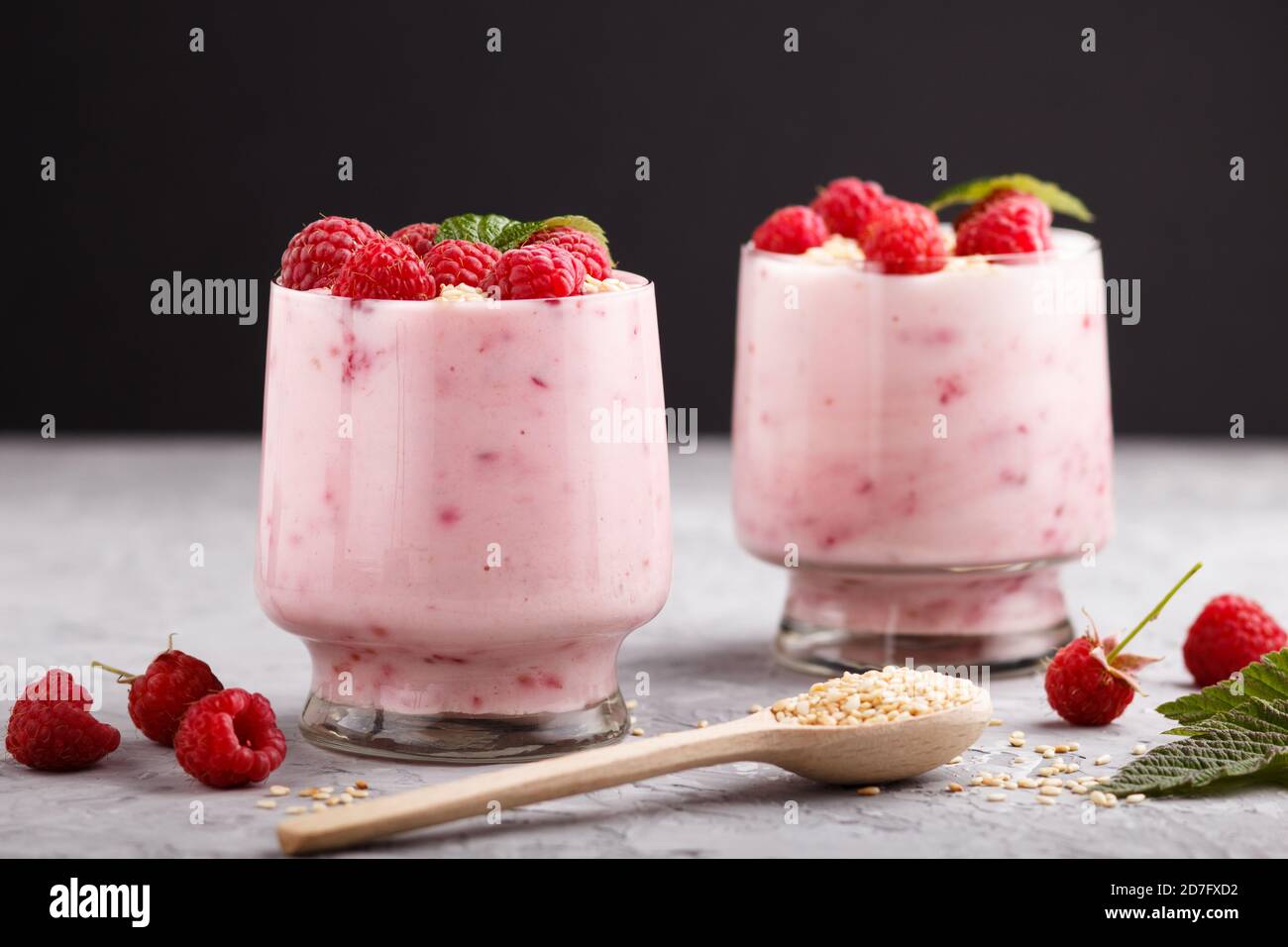 Joghurt mit Himbeere und Sesam in Glas und Holzlöffel auf grauem und schwarzem Hintergrund. Seitenansicht, Nahaufnahme. Stockfoto