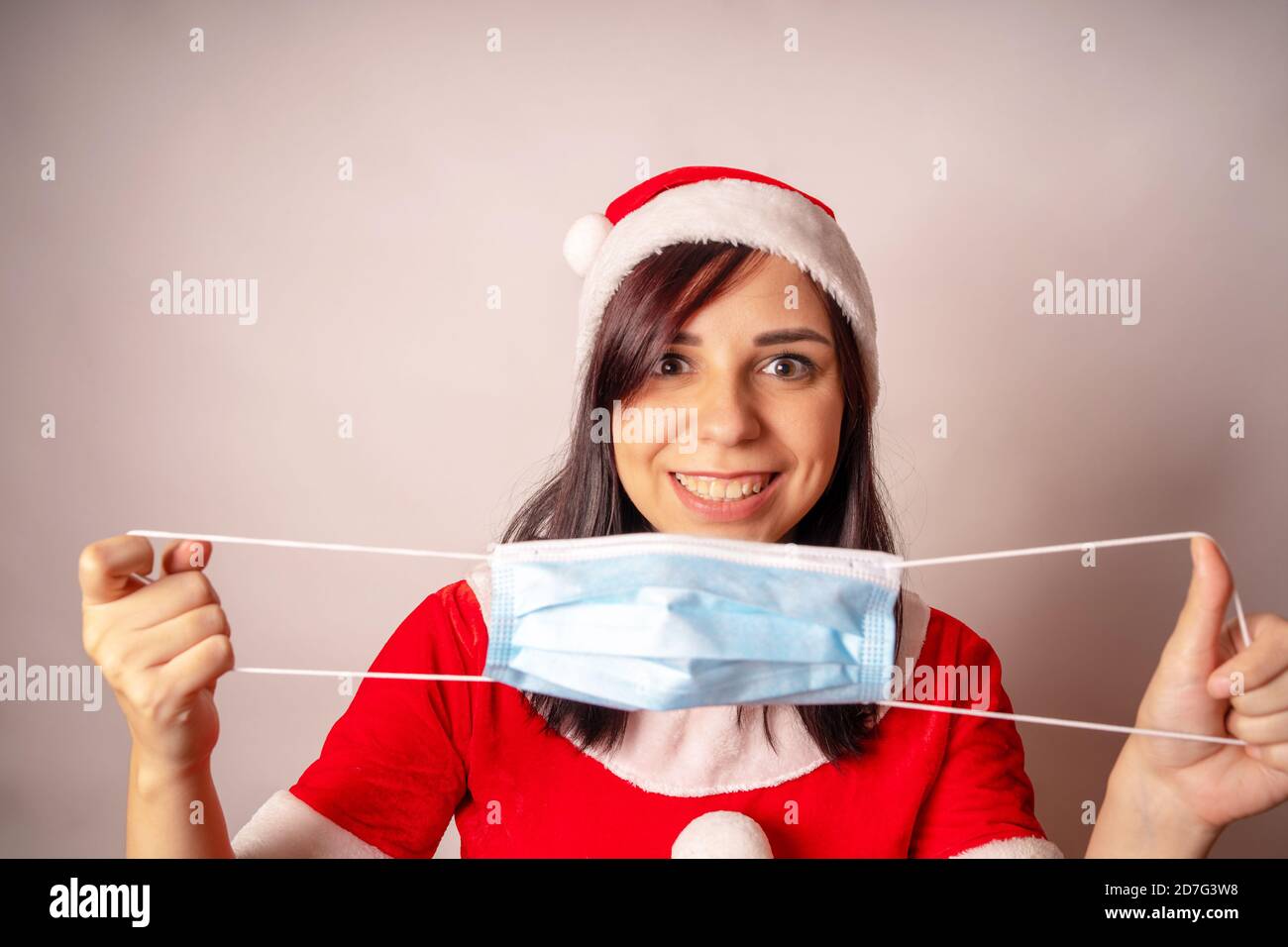 Frau im Weihnachtsmann Anzug mit medizinische Maske in den Händen auf grauem Hintergrund. Portrait der Brünette in Weihnachtsmütze mit Schutzmaske. Konzept von Stockfoto