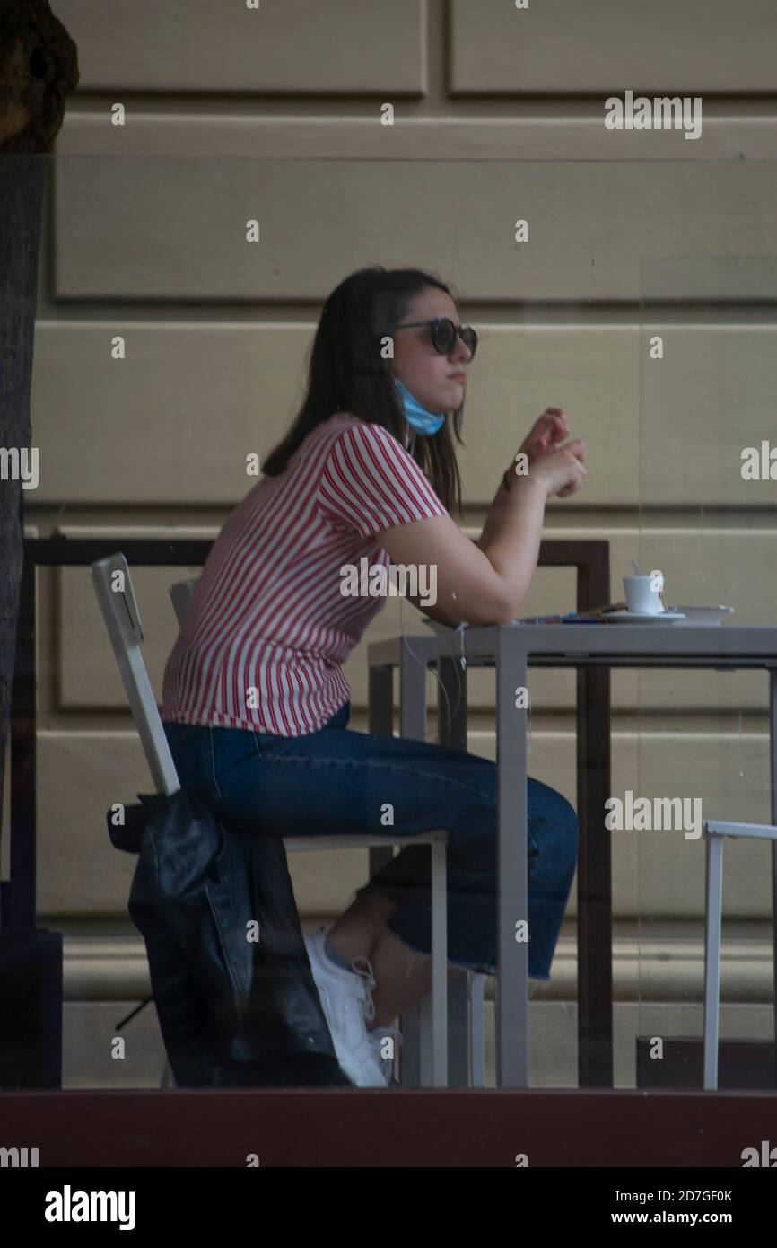 das italienische Mädchen sitzt allein, mit einer Maske unter dem Kinn, an einem Couchtisch, genießt einen Kaffee und reflektiert nachdenklich Stockfoto