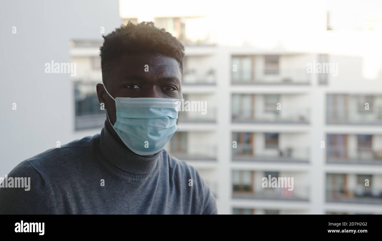 Schwarzer Mann mit medizinischer Maske auf dem Balkon und Blick auf die Kamera. Quarantäne und psychische Probleme. Hochwertige Fotos Stockfoto