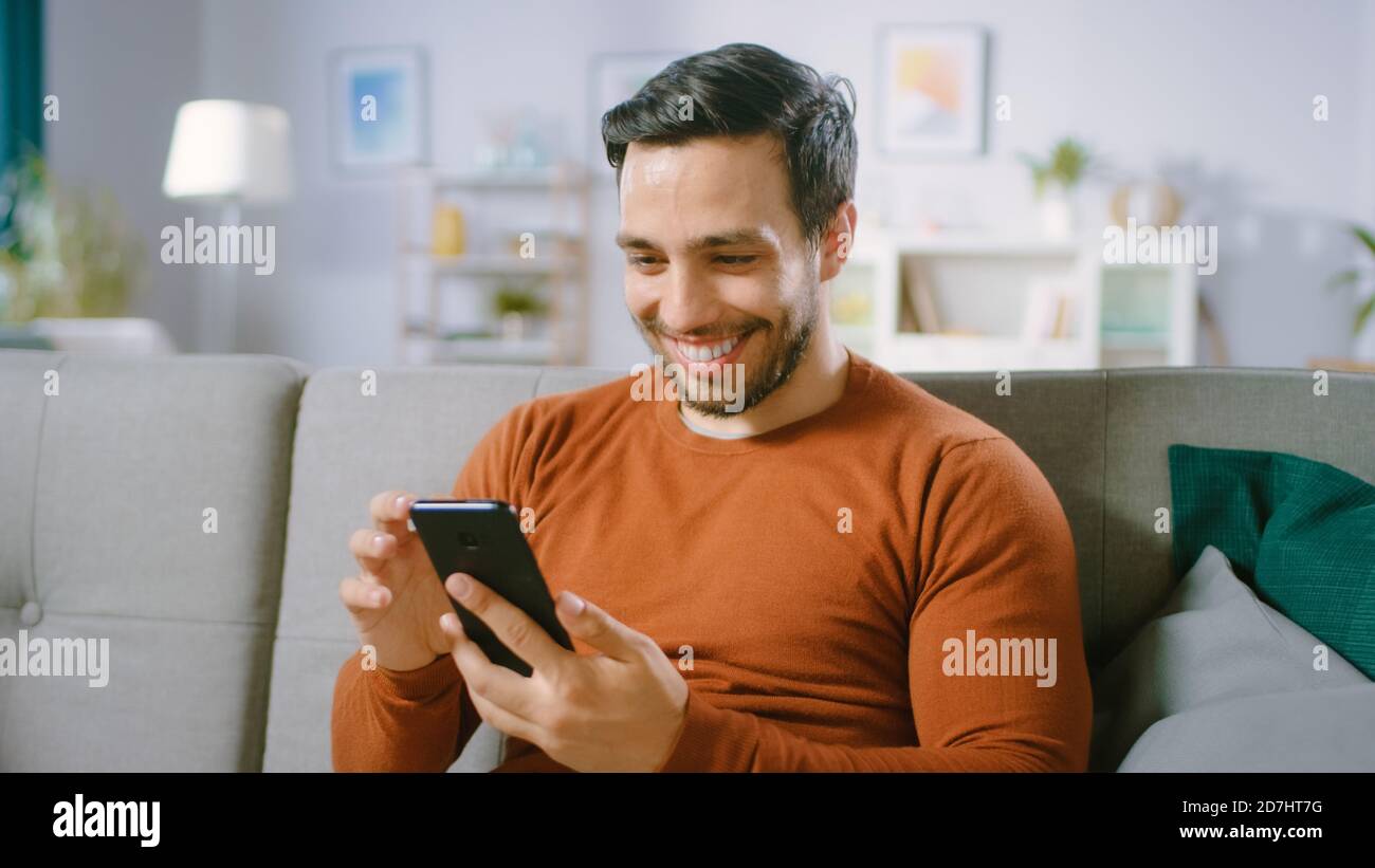 Happy Young man nutzt Smartphone, während er zu Hause auf einem Sofa sitzt. Der Mensch durchsucht das Internet, beobachtet Videos und nutzt soziale Netzwerke zu Hause. Stockfoto