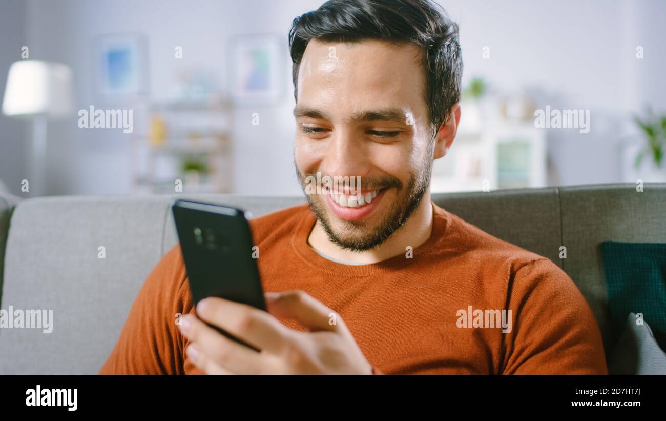 Happy Young man nutzt Smartphone, während er zu Hause auf einem Sofa sitzt. Der Mensch durchsucht das Internet, beobachtet Videos und nutzt soziale Netzwerke zu Hause. Stockfoto