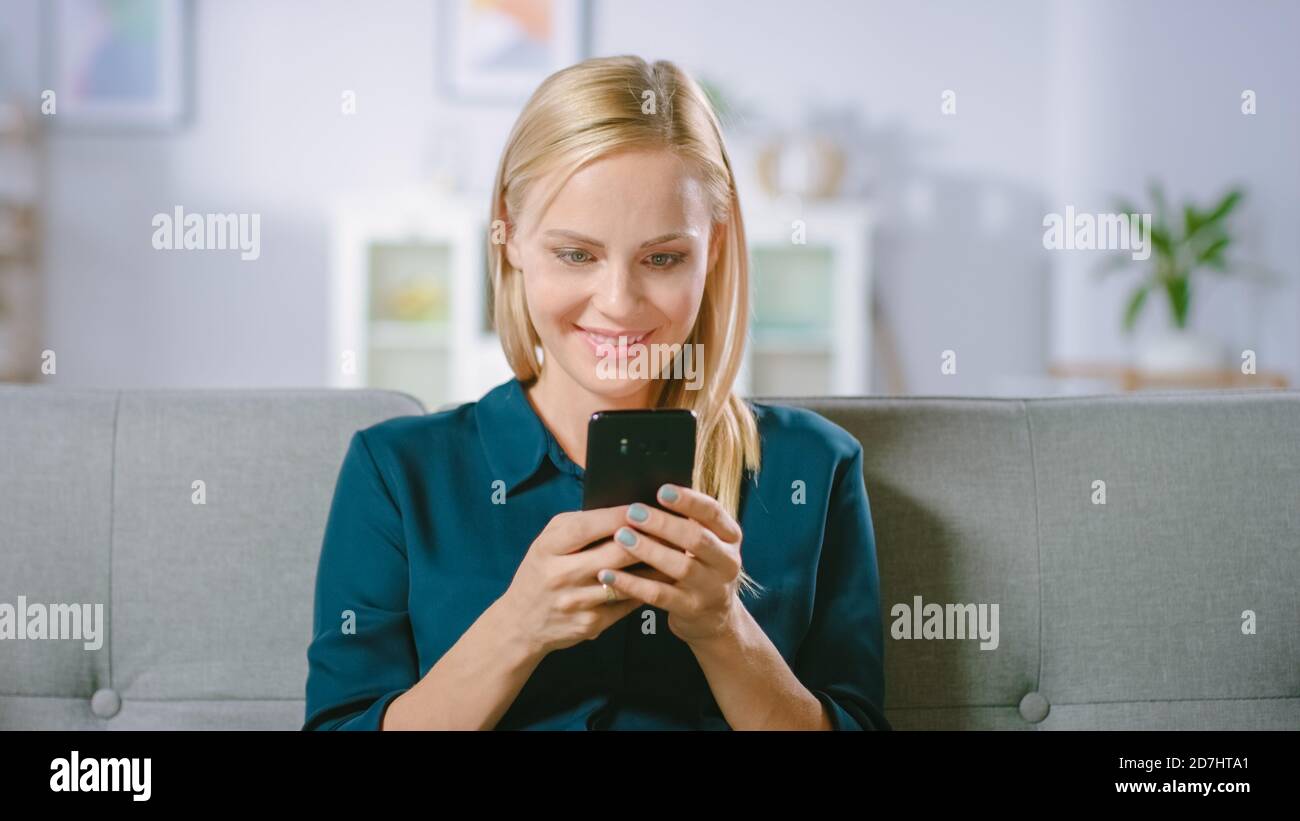 Schöne Blonde Frau nutzt Handy, während auf einem Sofa zu Hause sitzen. Smiling Happy Woman nutzt Smartphone für das Surfen im Internet, Social Stockfoto