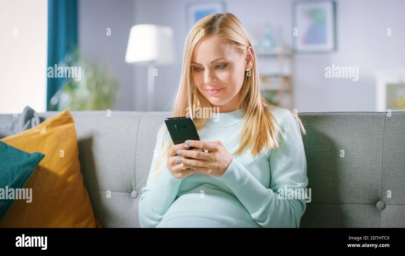 Schöne glücklich schwangere Frau auf einer Couch sitzt durchstösst Internet auf Smartphone. Future Mom macht Internet Shopping auf Smartphone von zu Hause. Stockfoto
