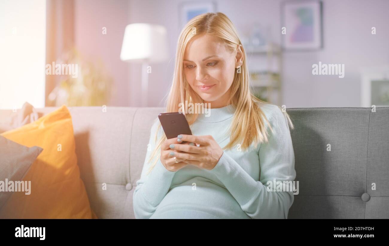 Schöne glücklich schwangere Frau auf einer Couch sitzt durchstösst Internet auf Smartphone. Future Mom macht Internet Shopping auf Smartphone von zu Hause Stockfoto