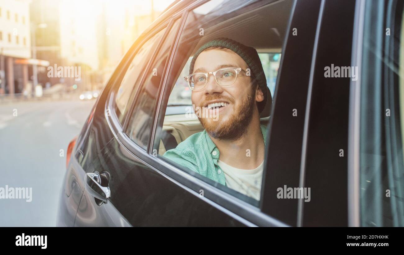 Stilvoller junger Mann fährt auf einem Beifahrer-Rücksitz eines Autos, schaut aus dem Fenster in Wonder. Großer sonniger Blick auf die Stadt spiegelt sich im Fenster. Schuss Gemacht Stockfoto