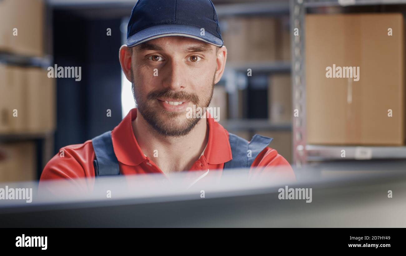 Porträt eines uniformierten Arbeiters, der einen Computer benutzt, während er an seinem Schreibtisch im Lager sitzt. Stockfoto