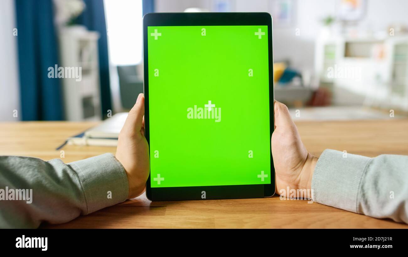 First Person Shot of man mit Green Mock-Up Screen Digital Tablet Computer im Portrait-Modus, während er an seinem Schreibtisch sitzt. Im Hintergrund Cosy Living Stockfoto