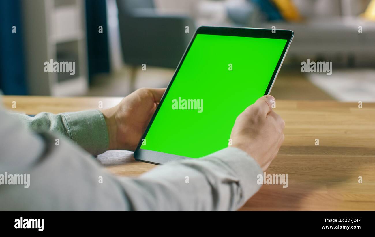 First Person Shot of man mit Handgesten auf Green Mock-Up Screen Digital Tablet Computer im Portrait-Modus, während er an seinem Schreibtisch sitzt. Im Stockfoto