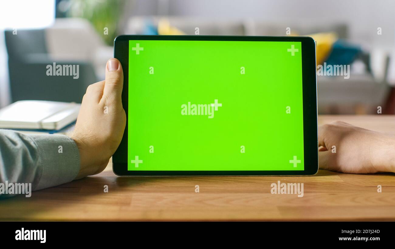 First Person Shot of a man mit Green Mock-up Screen Digital Tablet Computer im Querformat, während er an seinem Schreibtisch sitzt. Im Hintergrund gemütlich Stockfoto