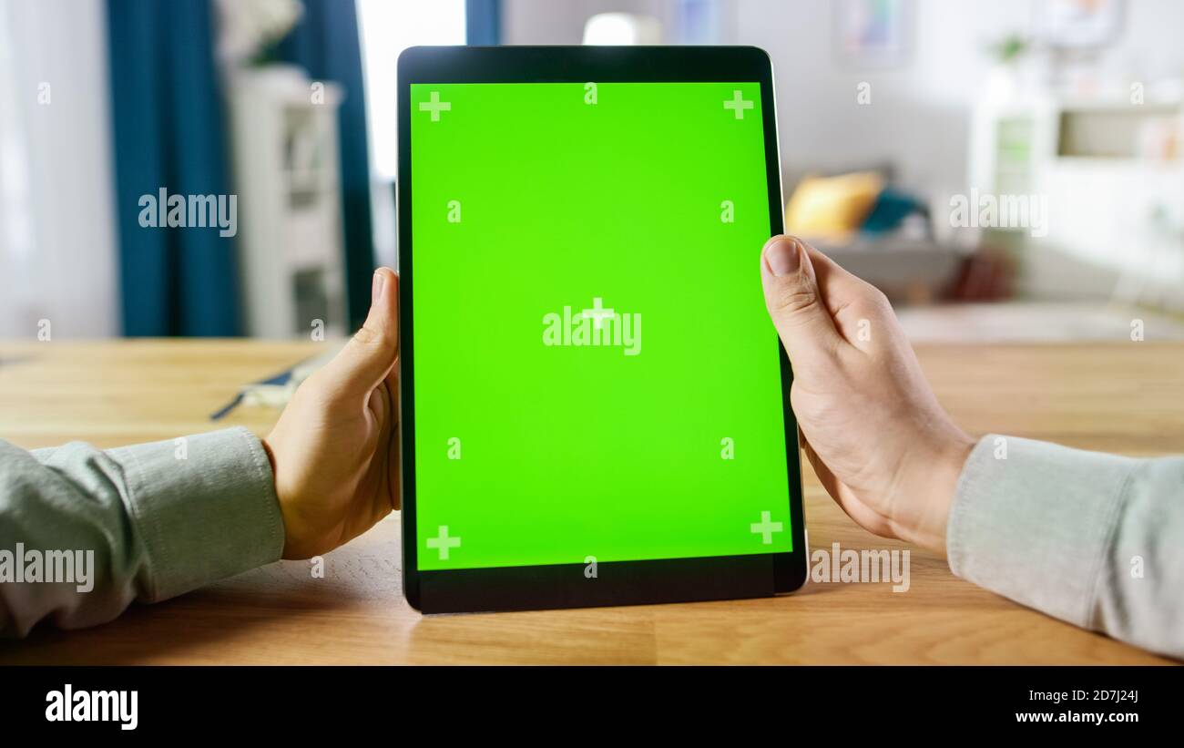 First Person Shot of a man mit Handgesten auf Green Mock-Up Screen Digital Tablet Computer im Portrait-Modus, während er an seinem Schreibtisch sitzt. Im Stockfoto