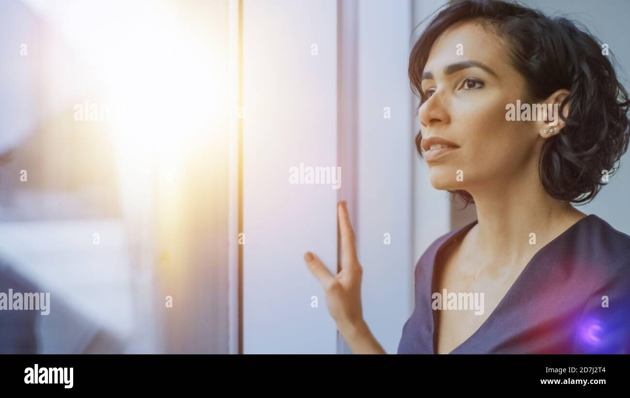 Porträt der schönen jungen Geschäftsfrau, die nachdenklich aus ihrem Bürofenster schaut. Selbstbewusste und attraktive hispanische Frau, die darüber nachdenkt Stockfoto