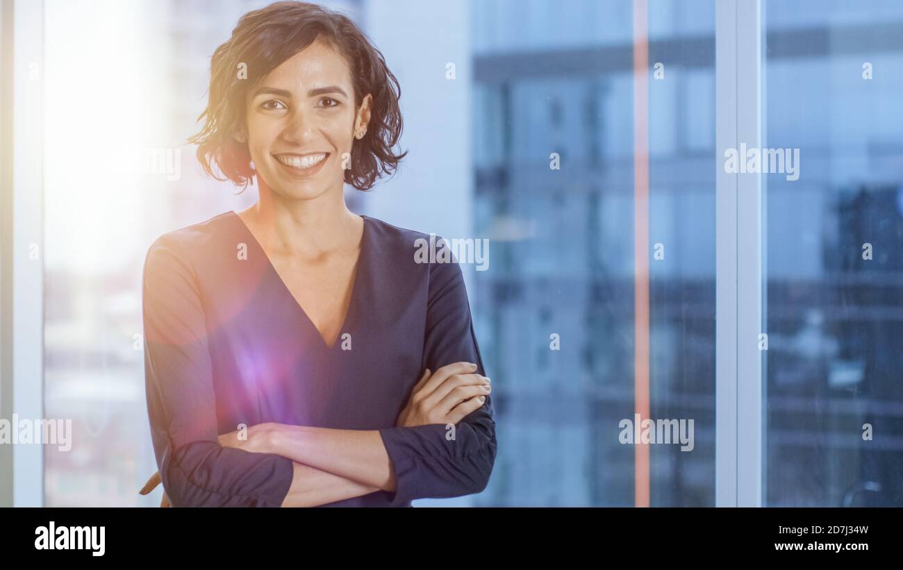 Porträt der erfolgreichen hispanischen Geschäftsfrau, die ihre Arme überquert und lächelt. Schöne weibliche Executive stehend in ihrem Büro. Sommertie Fackeln. Stockfoto