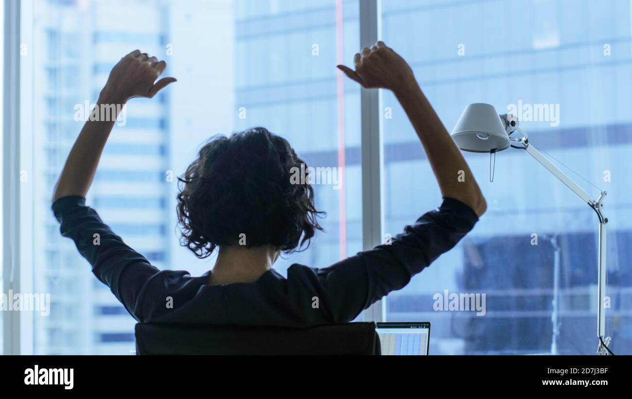 Rückansicht Aufnahme der schönen Geschäftsfrau, die an ihrem Schreibtisch sitzt und die Arme hebt in einer Feier einer erfolgreichen Jobförderung. Stockfoto