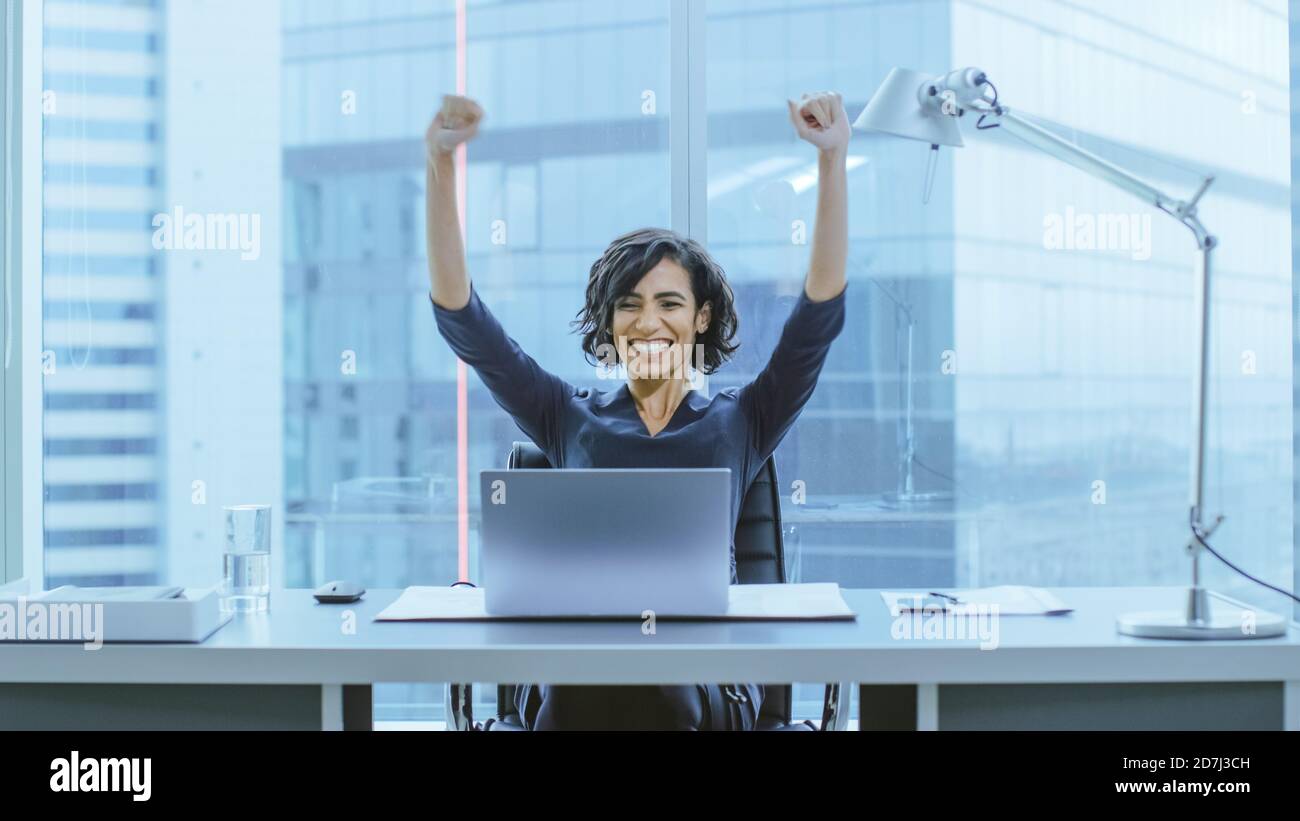 Aufnahme der schönen Geschäftsfrau, die an ihrem Schreibtisch sitzt und ihre Arme bei einer Feier einer erfolgreichen Jobförderung hebt. Stockfoto