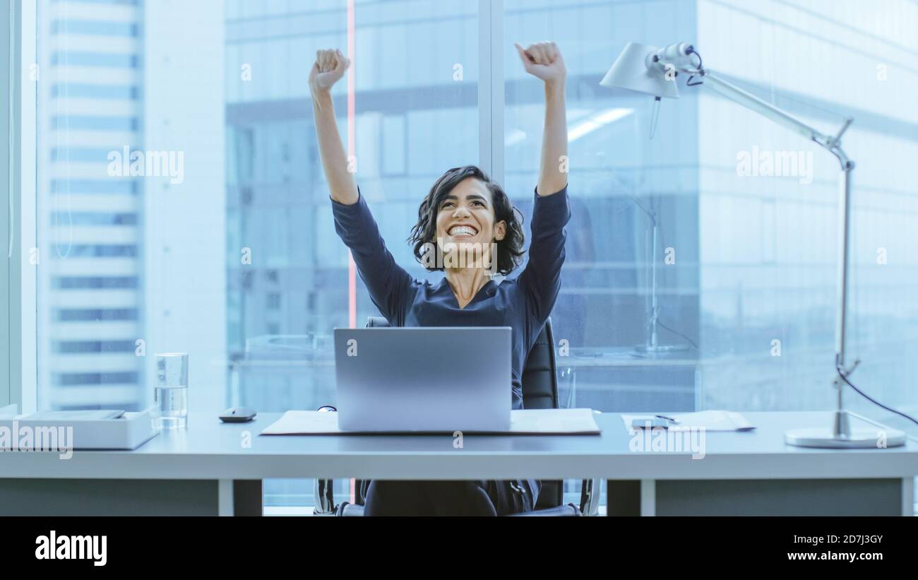 Aufnahme der schönen Geschäftsfrau, die an ihrem Schreibtisch sitzt und ihre Arme bei einer Feier einer erfolgreichen Jobförderung hebt. Stockfoto