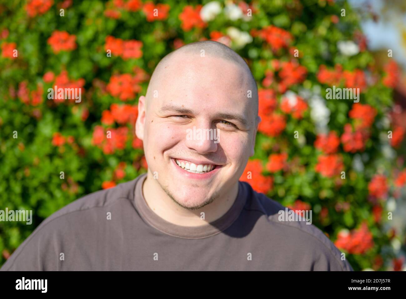 Aufsehenliches Porträt eines jungen Mannes, der die Augen zudreht Beim Lächeln im Freien in der Sonne vor einem Hintergrund von Bunte rote Blumen in der Garde Stockfoto