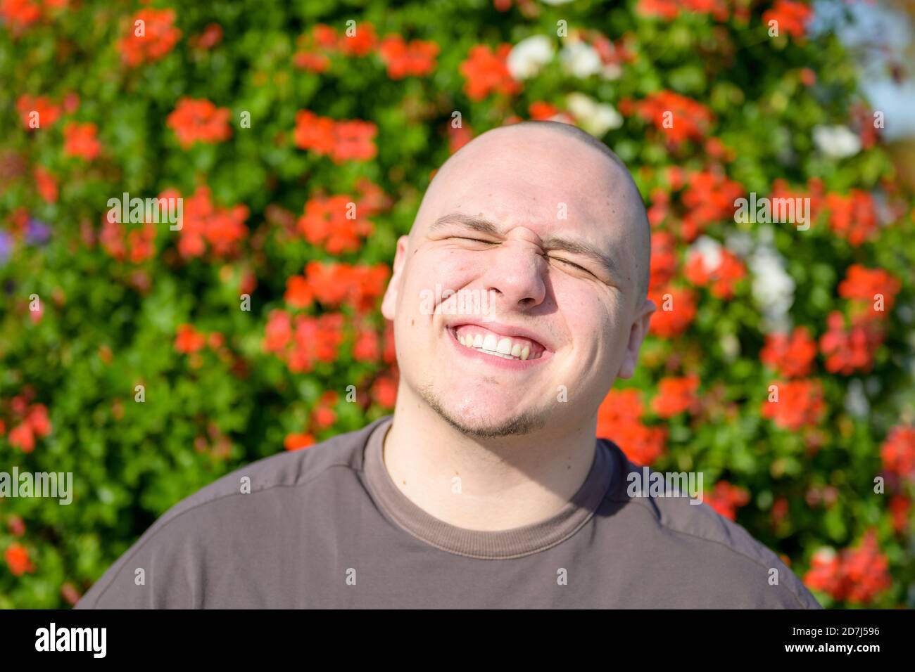Aufsehenliches Porträt eines jungen Mannes, der die Augen zudreht Beim Lächeln im Freien in der Sonne vor einem Hintergrund von Bunte rote Blumen in der Garde Stockfoto