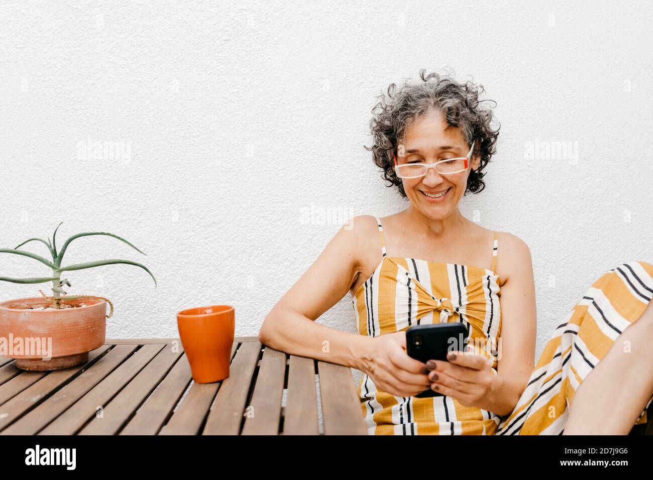 Lächelnde reife Frau mit kurzen Haaren mit Smartphone während Sitzen gegen weiße Wand im Hinterhof Stockfoto