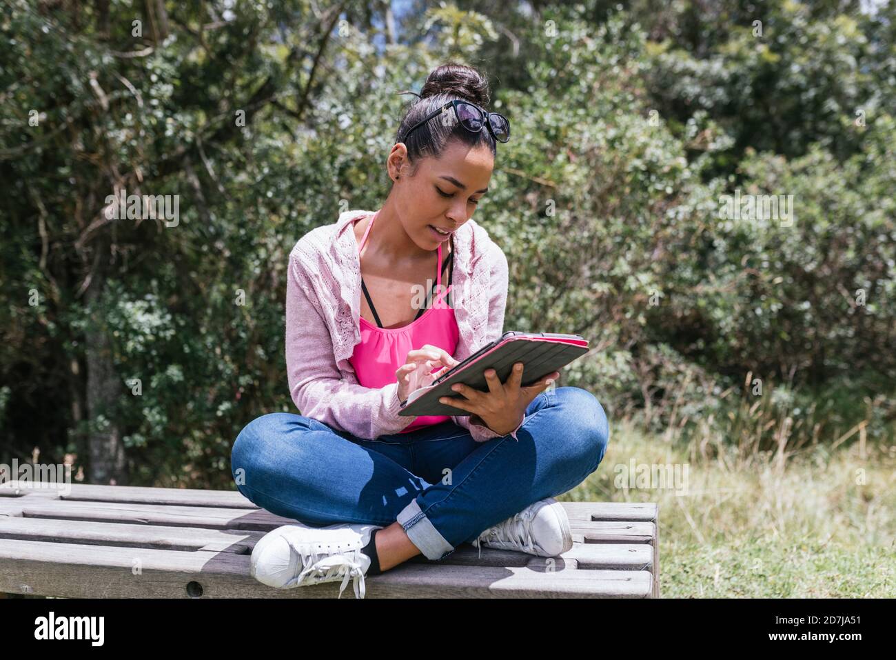 Junge Frau mit digitalen Tablet, während sie mit gekreuzten Beinen sitzt An sonnigen Tagen auf der Bank im Park Stockfoto