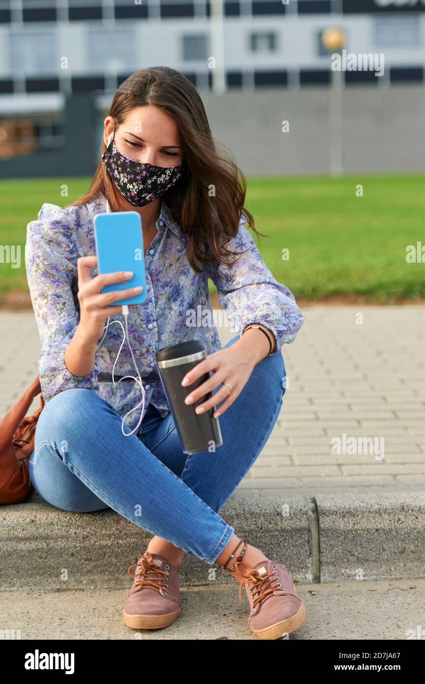 Junge Frau mit Gesichtsmaske mit Handy während des Sitzens Auf Fußweg in der Stadt Stockfoto