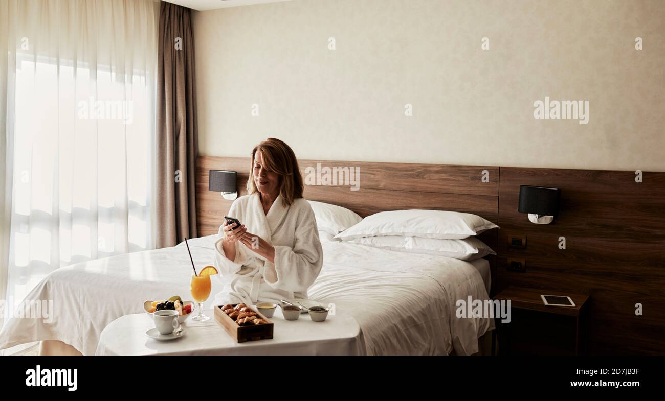 Blonde ältere Frau mit Smartphone während sitzen mit Frühstück Im Hotelzimmer Stockfoto