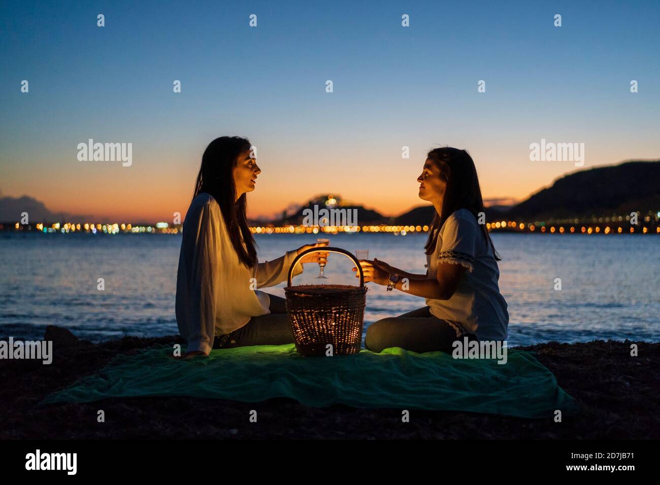 Schwestern mit Getränken reden, während sie am Strand auf der Decke sitzen Gegen klaren Himmel in der Dämmerung Stockfoto