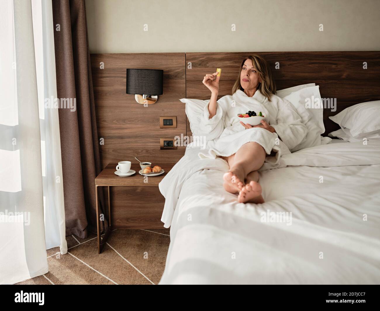 Ältere Frau, die beim Frühstück frisches Obst isst, während sie sich entspannt Bett und Blick auf das Hotelzimmer Stockfoto