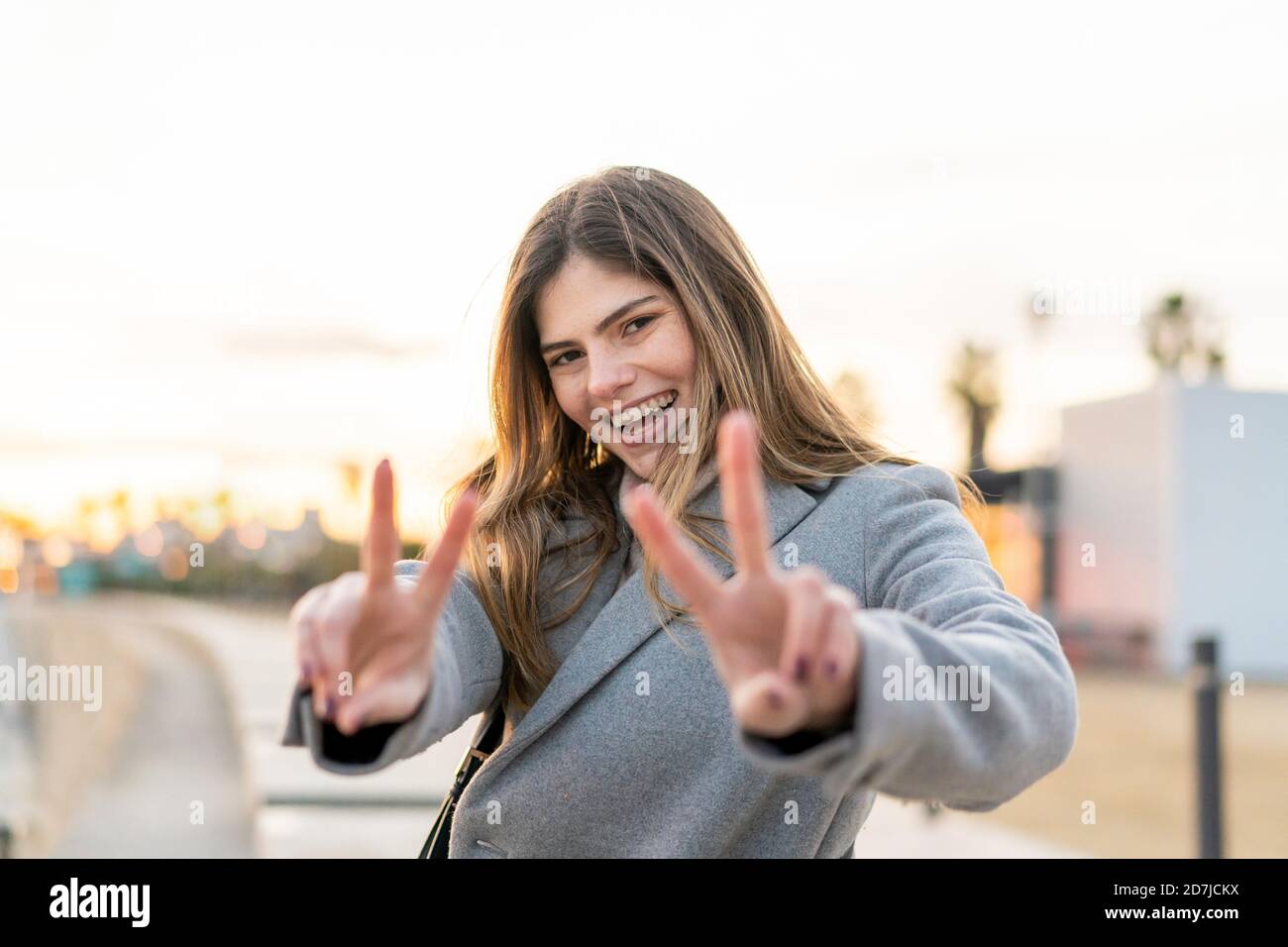 Glückliche Frau zeigt Friedenszeichen mit Fingern, während sie steht Strand bei Sonnenuntergang Stockfoto