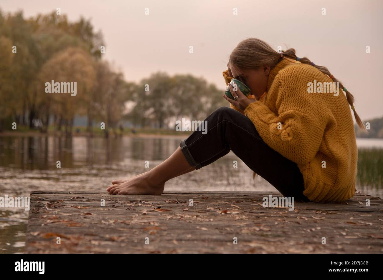 Ein barfuss Mädchen in einem gelben Strickpullover sitzt auf der Brücke über den See und trinkt aus einem grünen Becher. Stockfoto