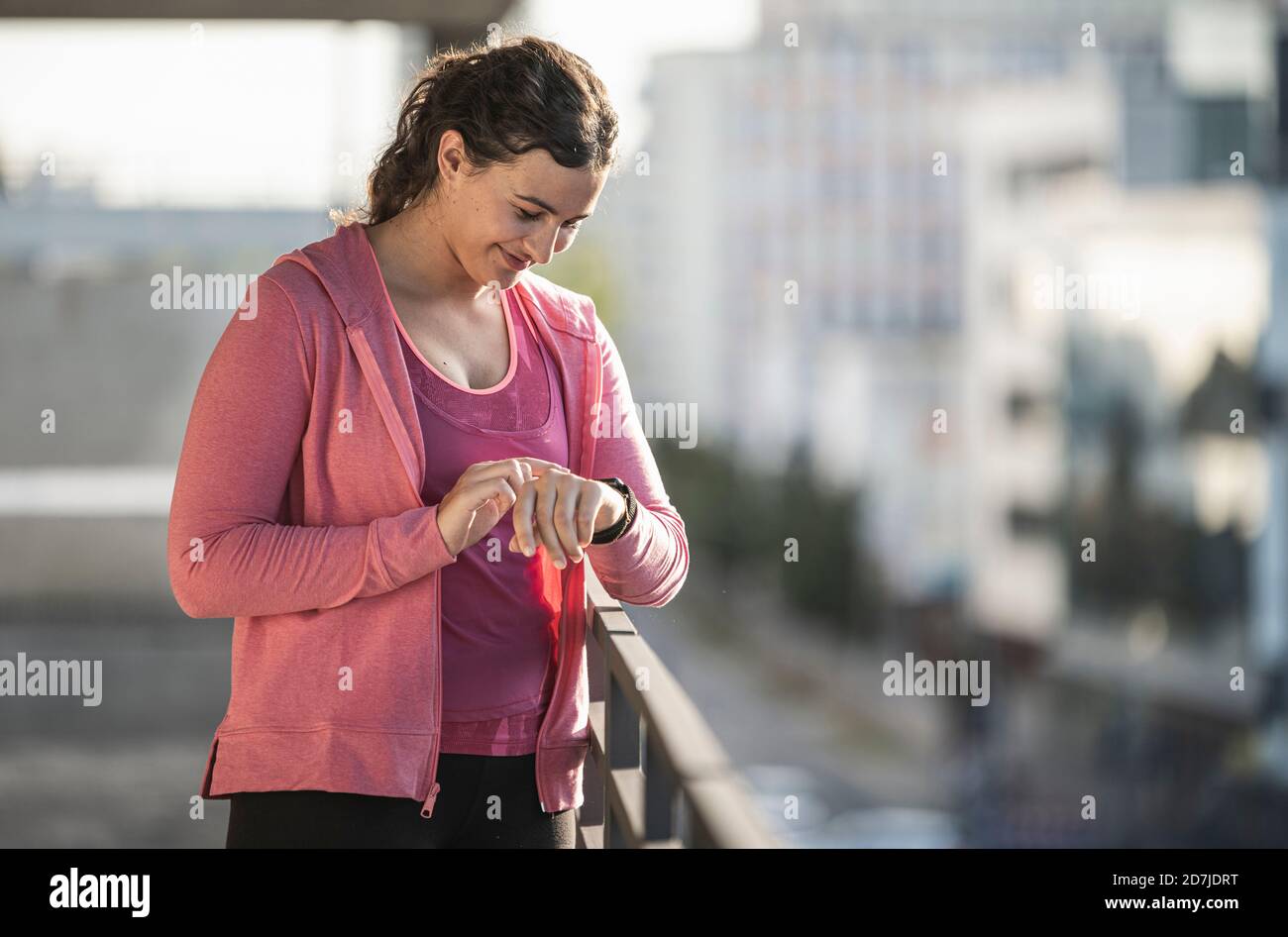 Lächelnde junge Frau, die Zeit überprüft, während sie beim Geländer steht Sonniger Tag Stockfoto