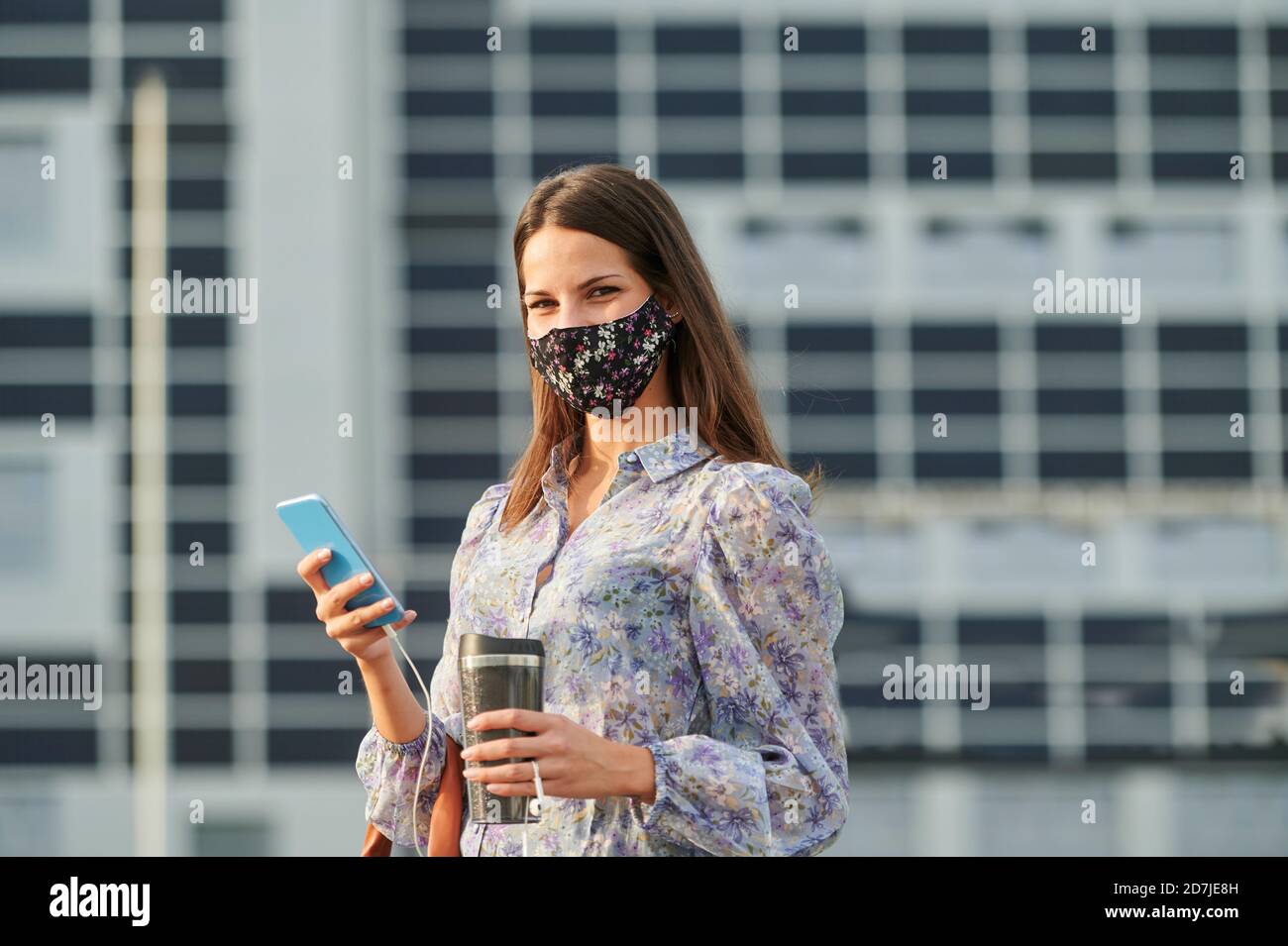 Junge Frau trägt Gesichtsmaske hält Thermoskanne während der Verwendung von mobilen Telefon in der Stadt Stockfoto