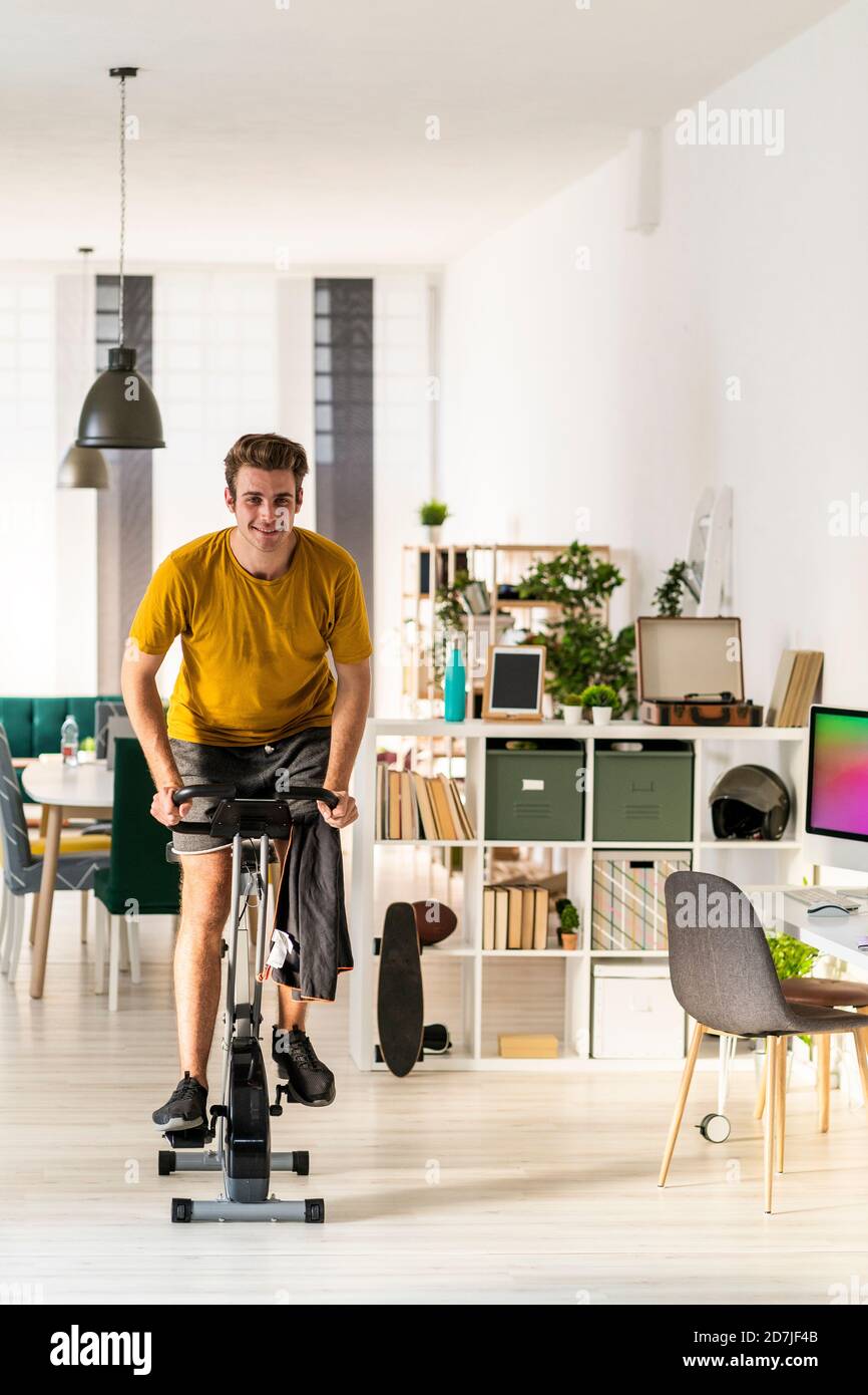 Lächelnder junger Mann, der zu Hause auf Trainingsgeräten radelt Stockfoto