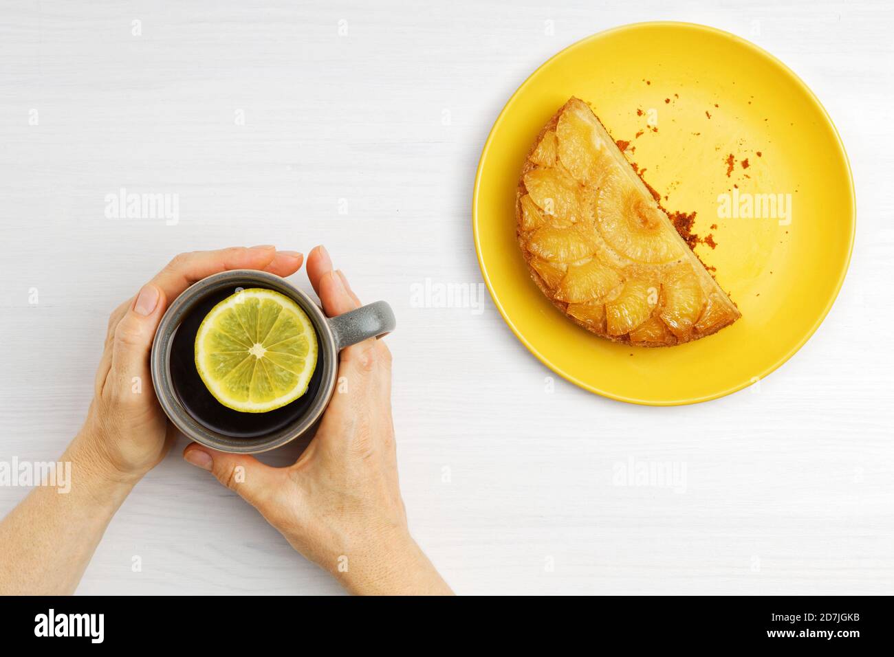 Weibliche Hände halten eine warme Tasse Tee mit einer Scheibe Zitrone und Stück Ananas auf dem Kopf Kuchen. Draufsicht. Stockfoto