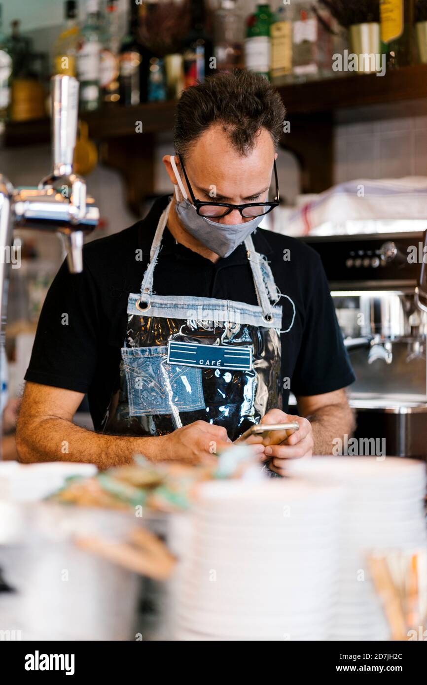 Barista in Schutzmaske mit Smartphone bei Cafe während der COVID-19 Stockfoto