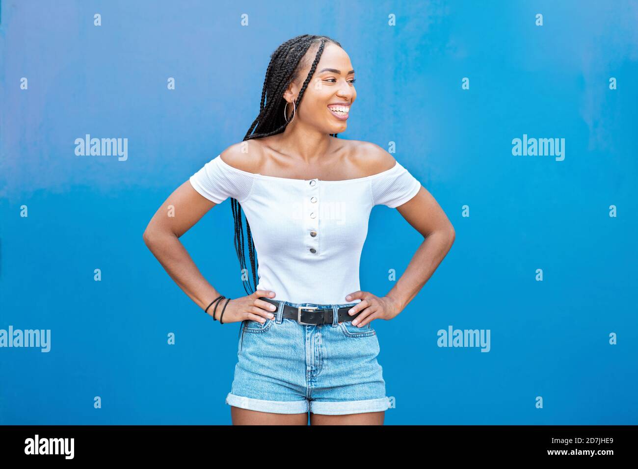 Lächelnde Frau mit der Hand an der Hüfte, die gegen die blaue Wand steht Stockfoto