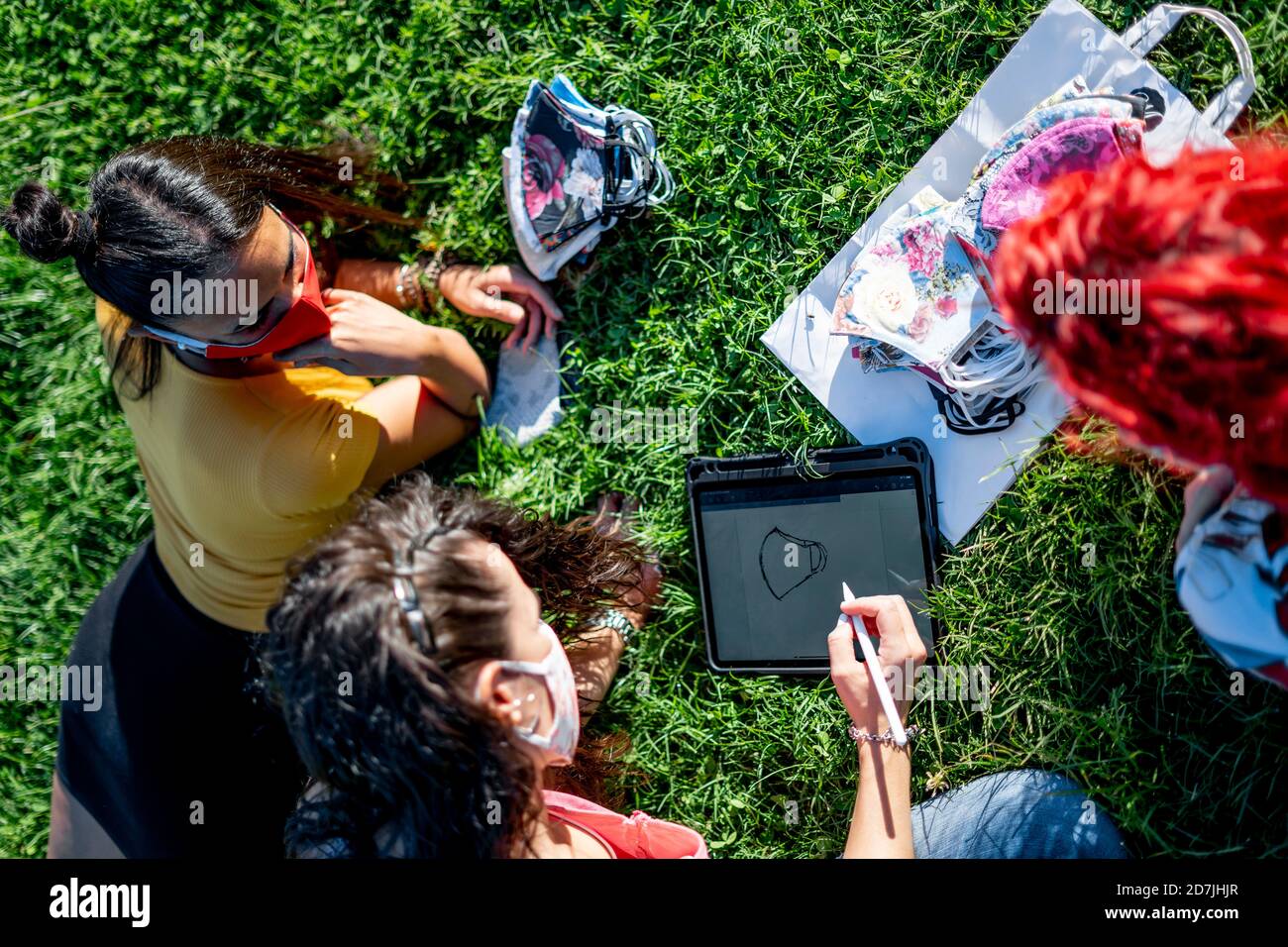 High-Angle-Ansicht von Freunden entwerfen schützende Gesichtsmaske mit Digitales Tablet, während Sie auf dem Rasen sitzen Stockfoto