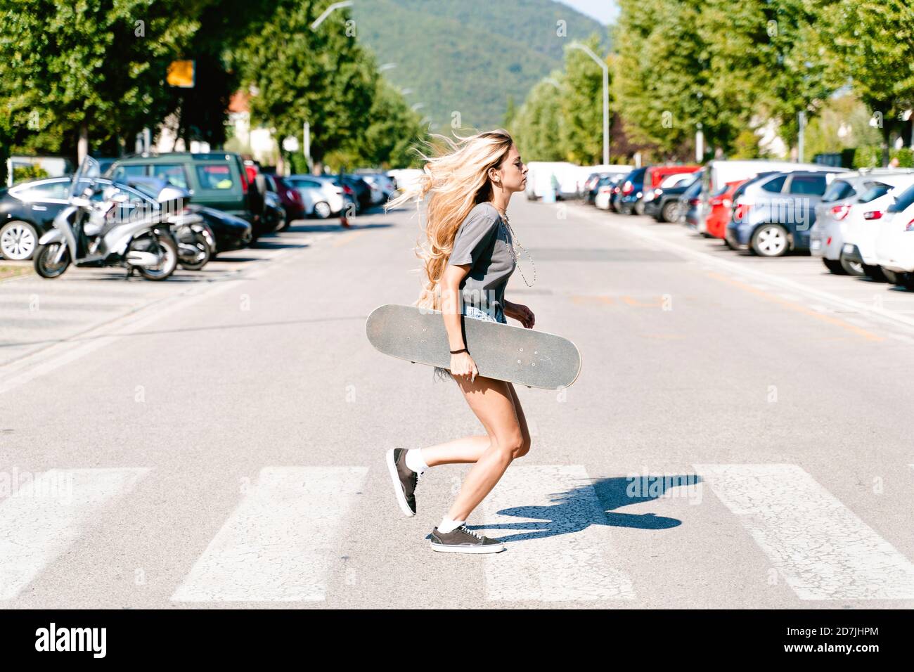 Junge blonde Frau hält Skateboard, während sie auf Zebrastreifen läuft In der Stadt Stockfoto